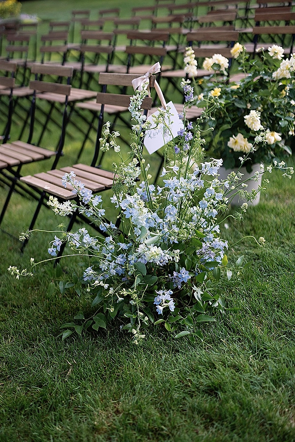 Ceremony aisle decor with blue delphinium and garden roses on grass beside wooden folding chairs and a reserved seat sign with ribbon bow