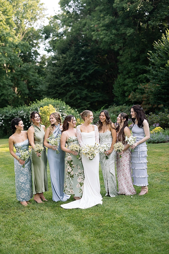Bridesmaids group photo with bride holding white flower and baby’s breath bouquets, smiling together on a garden lawn with greenery