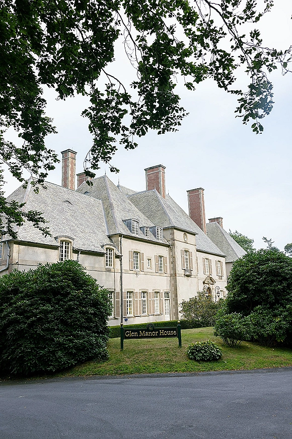 Wedding venue exterior of a manor house with slate roof and brick chimneys, venue sign by hedges, framed by trees under overcast sky