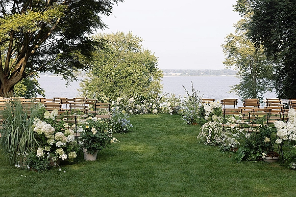 Outdoor ceremony setup with wood bench seating and white hydrangea aisle flowers on a lawn overlooking a calm lake and trees