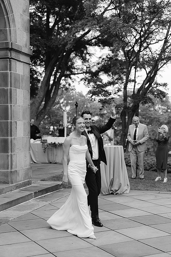 Wedding recessional as newlyweds walking away hand in hand under a stone archway, groom waving beside high-top cocktail tables and guests