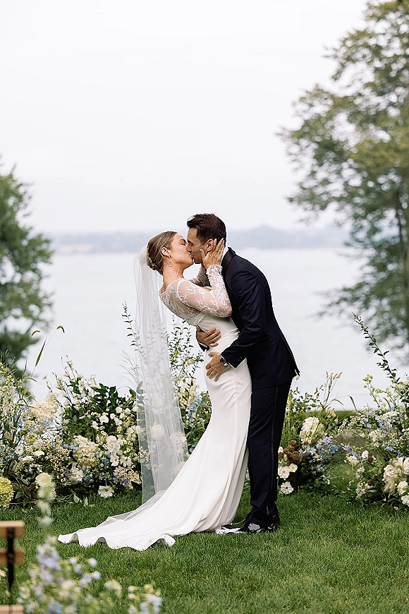 Wedding kiss portrait of bride and groom kissing, her cathedral veil flowing as she cups his face by a calm lakeside lawn and trees