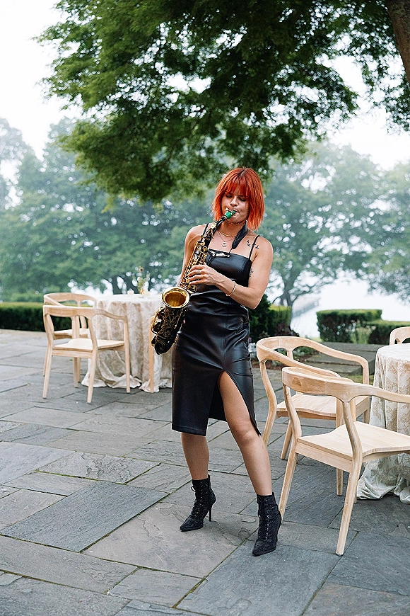 Wedding musician saxophonist at wedding in a black dress playing on a stone patio beside round tables, trees, and distant water view