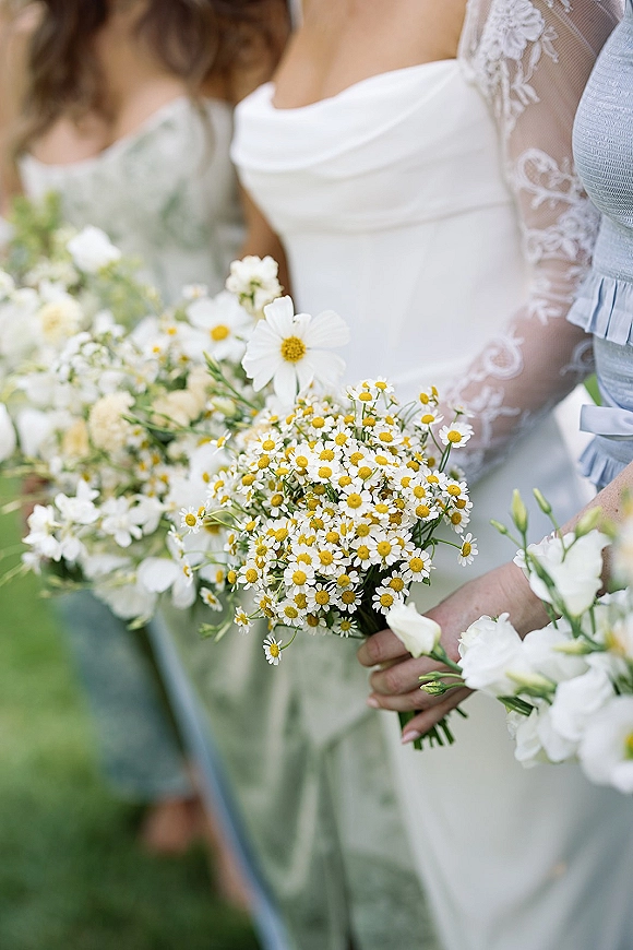 Bridal bouquet of daisies and chamomile held close by a bride in a lace sleeve gown, with bridesmaids in sage dresses amid greenery