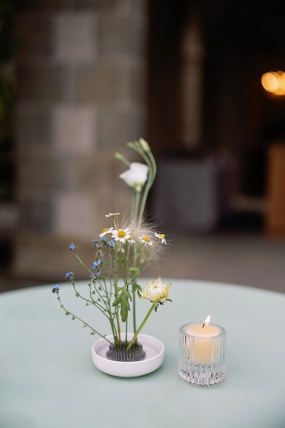 Reception table centerpiece with a minimal floral centerpiece of chamomile daisies and blue wildflowers in a bowl beside a glass votive candle, set near a stone wall doorway