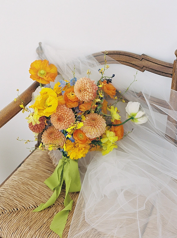 Bridal bouquet with bright wedding bouquet blooms tied with a green ribbon, resting on a woven chair seat against a white wall
