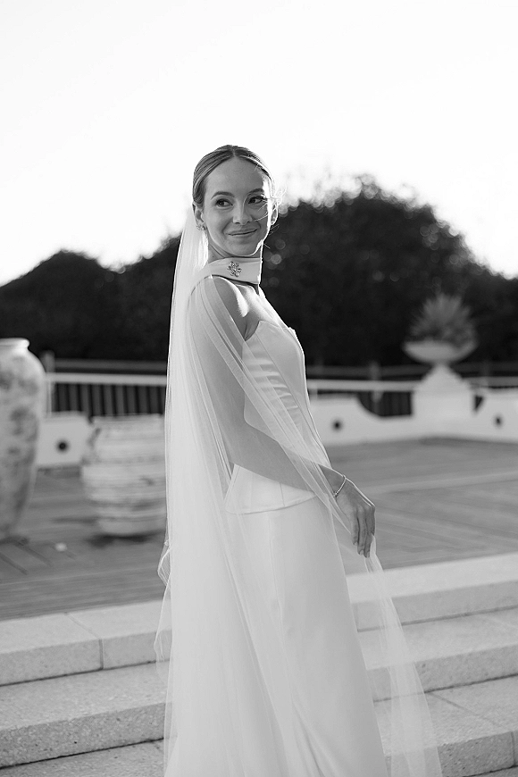 Bridal portrait in black and white of a bride looking over her shoulder in a high-neck gown with long veil on stone terrace steps