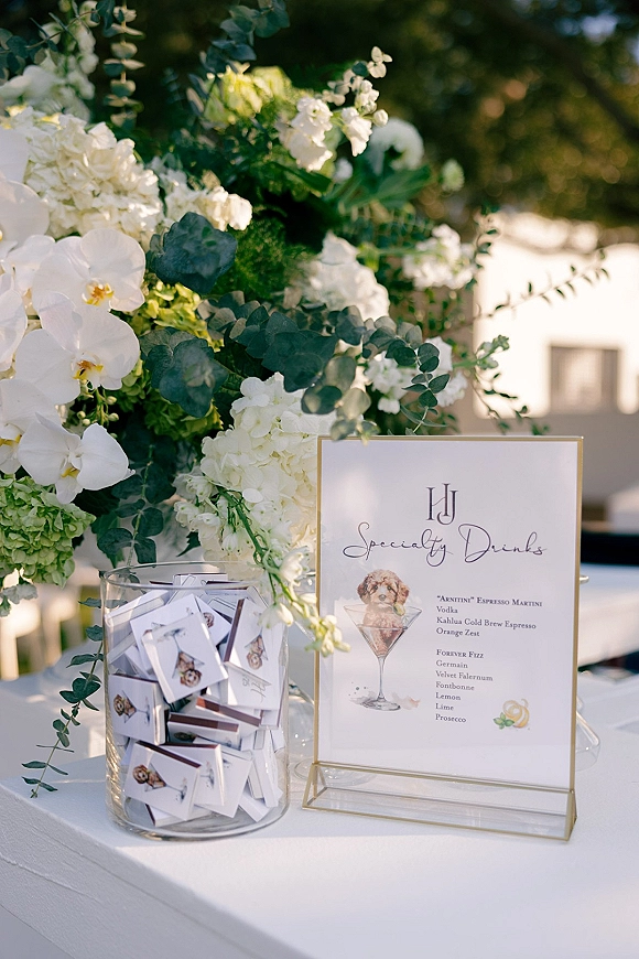 Wedding bar signage in a gold frame with specialty drinks wedding sign beside white orchids, hydrangeas, and eucalyptus on linen outdoors