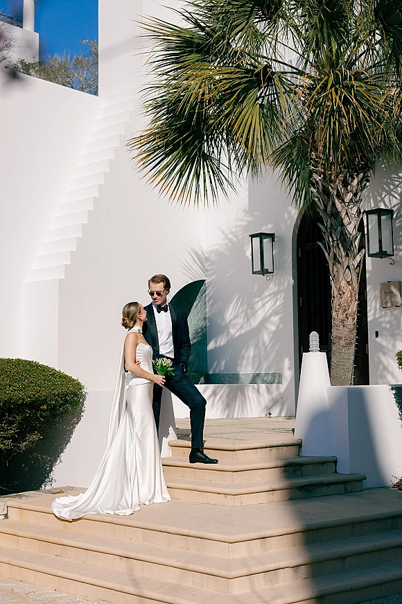 Couple portrait of bride and groom, bride in satin dress with long veil and bouquet, groom in tuxedo with sunglasses on stucco steps.