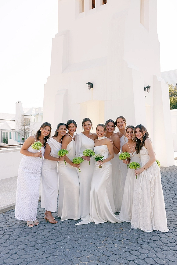 Bridesmaids portrait of women in white dresses holding green hydrangea bouquets, smiling in a paved courtyard by a white building lanterns