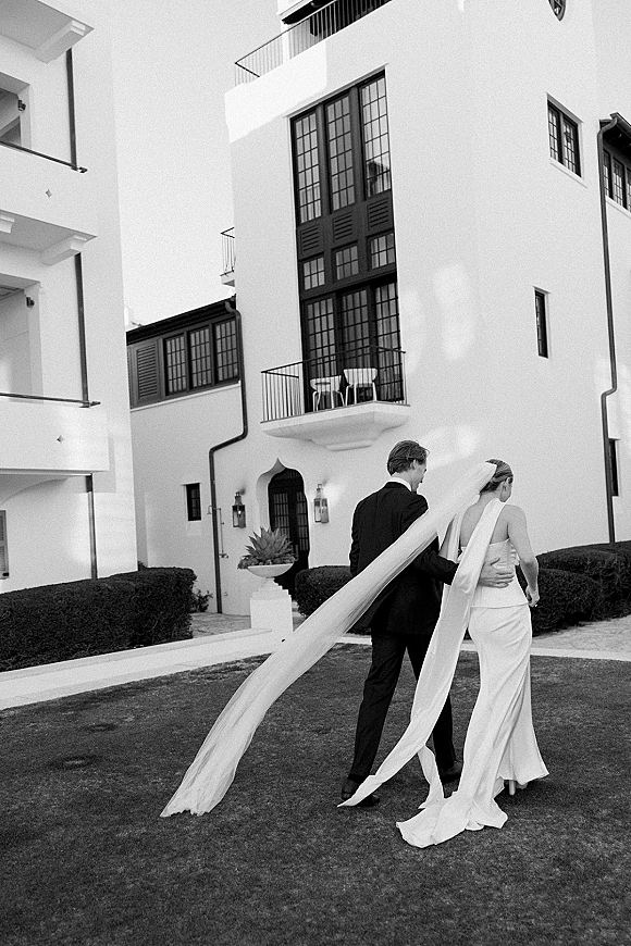Couple portrait of bride and groom walking away, groom arm around her as a long wedding veil train flows by a white villa backdrop
