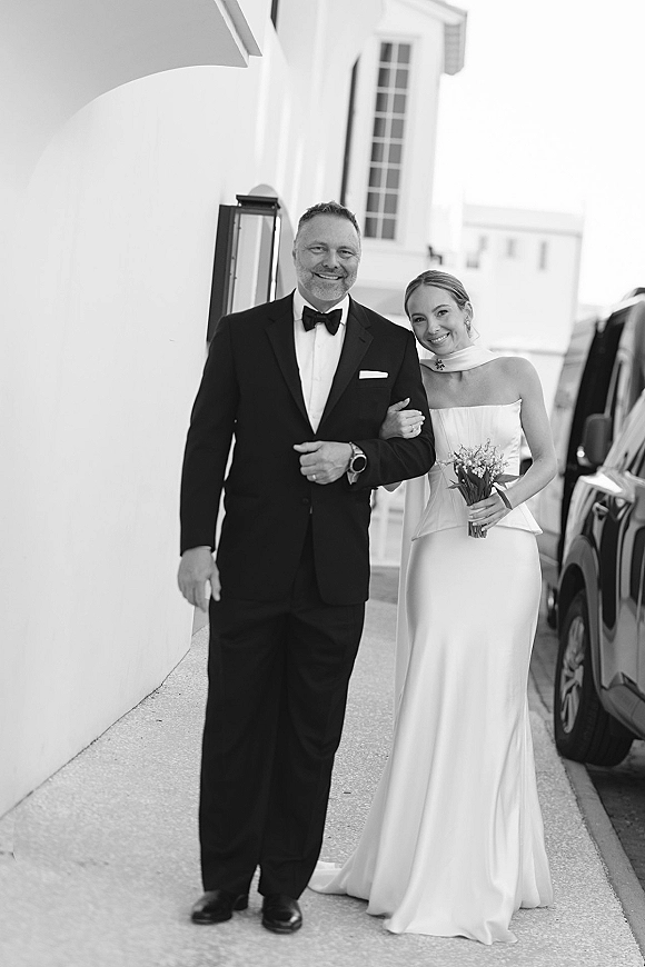 Bride with father walking arm in arm, she holds a bouquet in a strapless dress, outside a white building with lantern and parked car