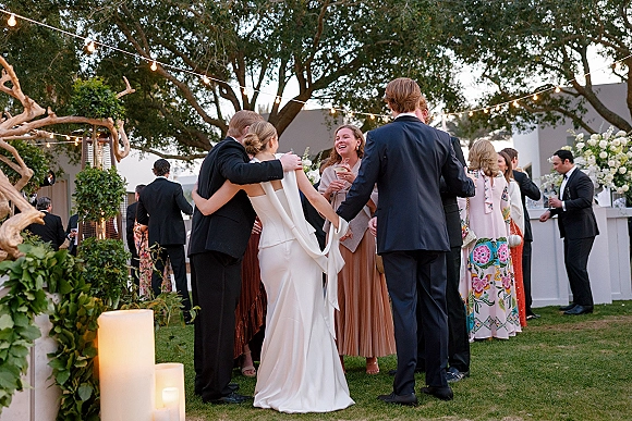 Wedding cocktail hour with guests mingling under string lights by an outdoor bar with pillar candles, greenery, and florals on a lawn by trees