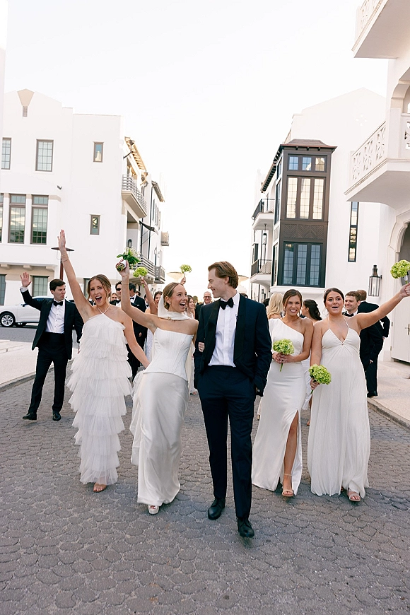Wedding party photo of bride and groom with bridesmaids walking and cheering on a cobblestone street lined with white buildings and balconies