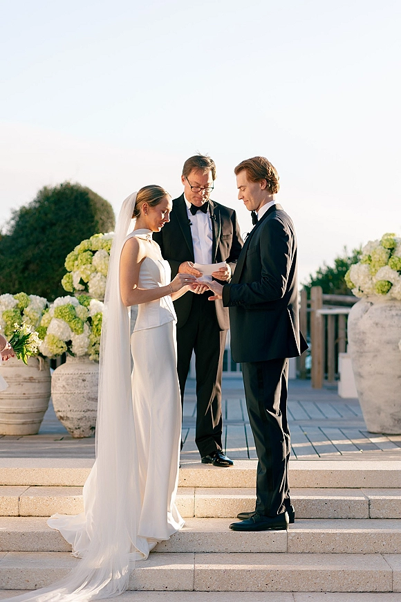 Wedding vows as bride and groom hold hands, her long veil and bouquet flowing on outdoor steps with hydrangea planters nearby