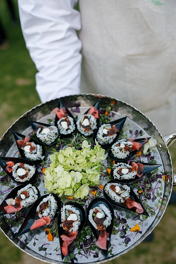 Wedding cocktail appetizers with sushi rolls in black cones on a silver tray, garnished with microgreens and ginger, beside hydrangeas on a lawn