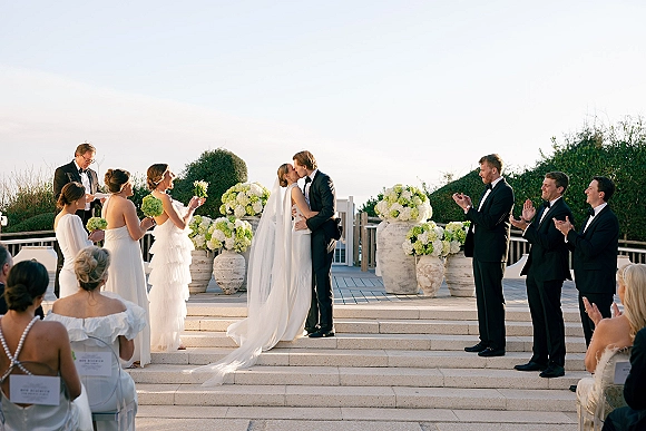 Ceremony kiss as bride in long veil and groom in tuxedo embrace on stone steps, flanked by bridesmaids and floral urns on terrace