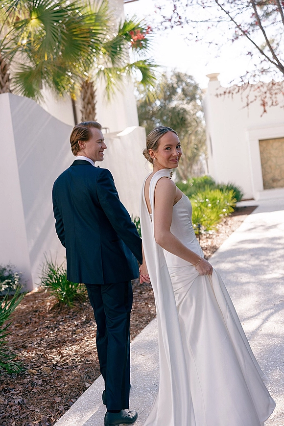 Couple portrait of bride and groom walking hand in hand, bride looking back in a satin dress and cape by palm trees and white stucco building