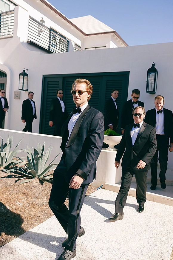 Groomsmen portrait with black tuxedos and sunglasses, standing on a concrete walkway by a white stucco building with black shutters