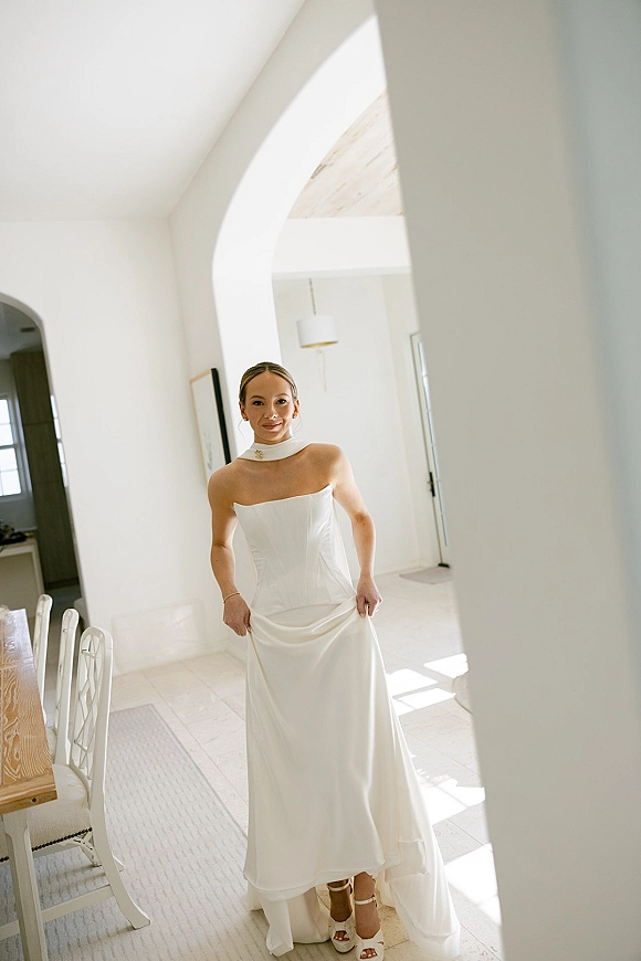 Bridal portrait of a bride in a strapless wedding dress with a neck scarf, standing by a white arched doorway in soft window light