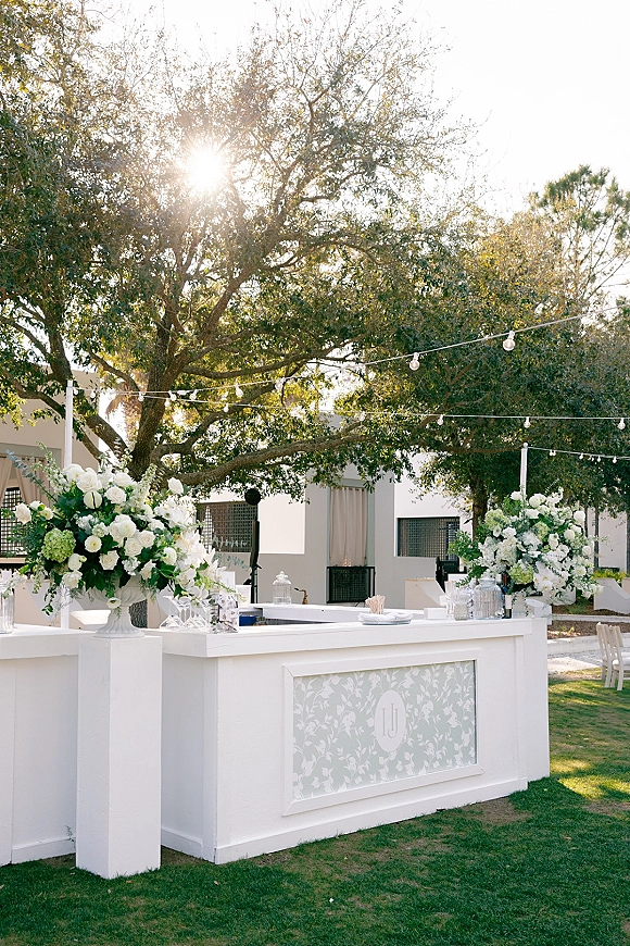 Wedding bar setup with a white bar, lush white and green florals, and glass drink dispensers under string lights on a sunny lawn