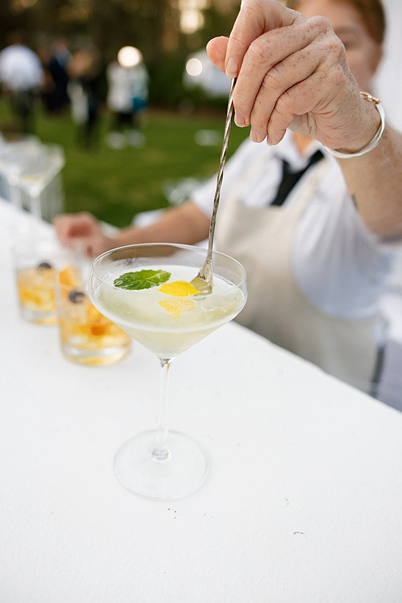 Wedding cocktail in a coupe glass with lemon peel and mint garnish beside a rocks glass with citrus and ice on an outdoor lawn bar setup