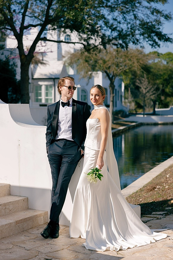 Couple portrait of bride in a strapless dress with long veil and bouquet beside groom in black tuxedo and sunglasses by a canal and white villa