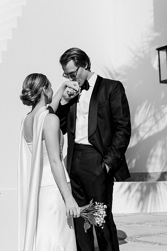 Wedding couple portrait of groom kissing bride’s hand as they hold hands, both in sunglasses by a stucco wall with tree shadows