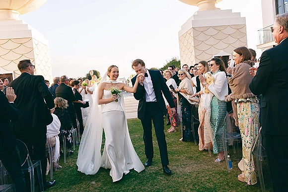 Wedding recessional as newlyweds walk down the aisle, groom kissing bride’s hand while guests cheer on a lawn by white columns