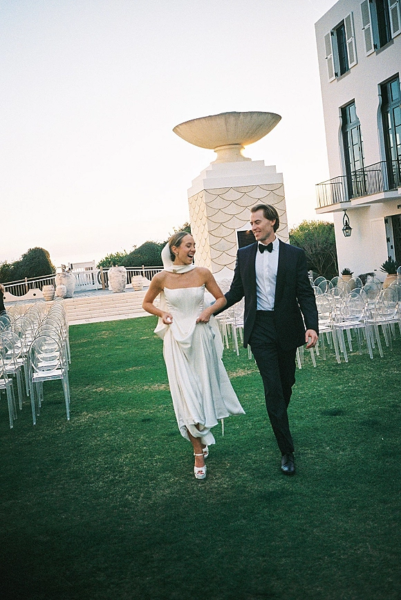 Wedding couple portrait of bride and groom walking, bride laughing in strapless dress and veil, groom in tuxedo before white facade lawn seating