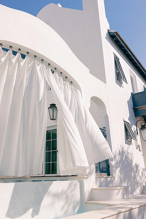 Wedding venue exterior with white drapery curtains framing an arched entrance on a white stucco building beside a pool under blue sky