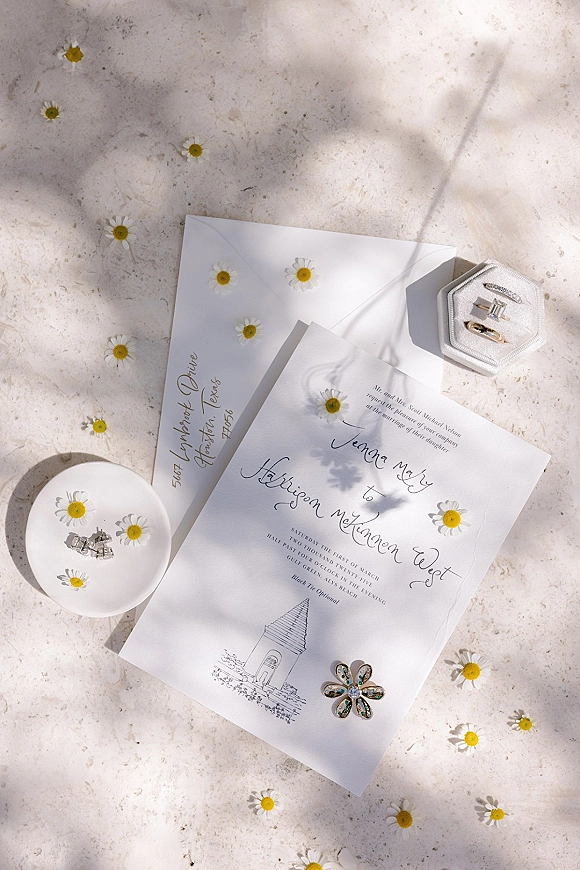 Invitation suite wedding invitation flat lay with calligraphy card, envelopes, daisies, ring box and jewelry on a light stone surface with shadows