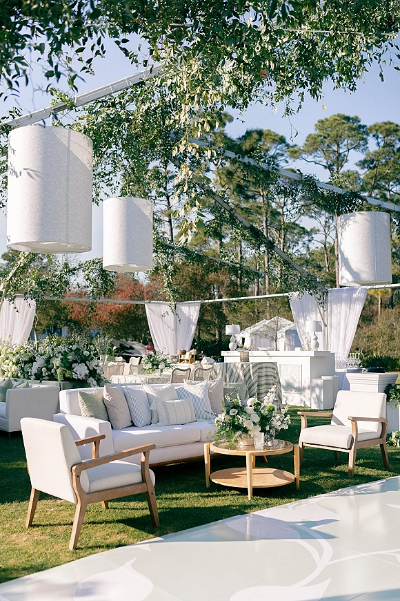 Wedding lounge area with white sofa and armchairs around a round coffee table, accented by greenery garlands and hanging lanterns under a tent