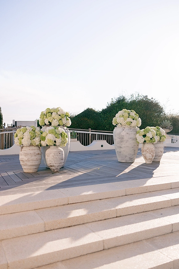 Ceremony altar decor with wedding altar flowers in large stone urns, featuring white and green hydrangeas on a wood deck by hedges