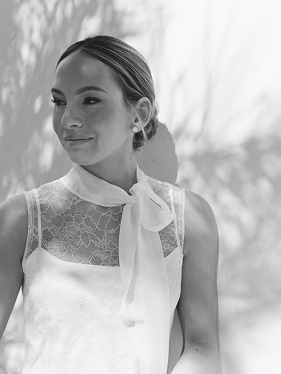 Bridal portrait of a bride looking away in a lace high neck dress with a bow, low bun, against a white wall with tree shadows