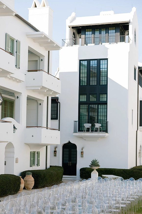 Outdoor ceremony setup with clear acrylic ceremony chairs in neat rows along a stone walkway, set against a white stucco facade and blue sky