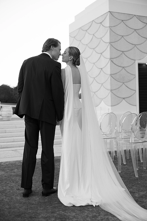 Couple portrait of bride and groom kissing, her long veil and cape flowing on terrace steps by a modern building facade