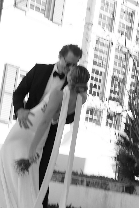 Wedding kiss portrait of groom dipping the bride for a kiss, her long veil flowing with bouquet, in black and white by a building facade