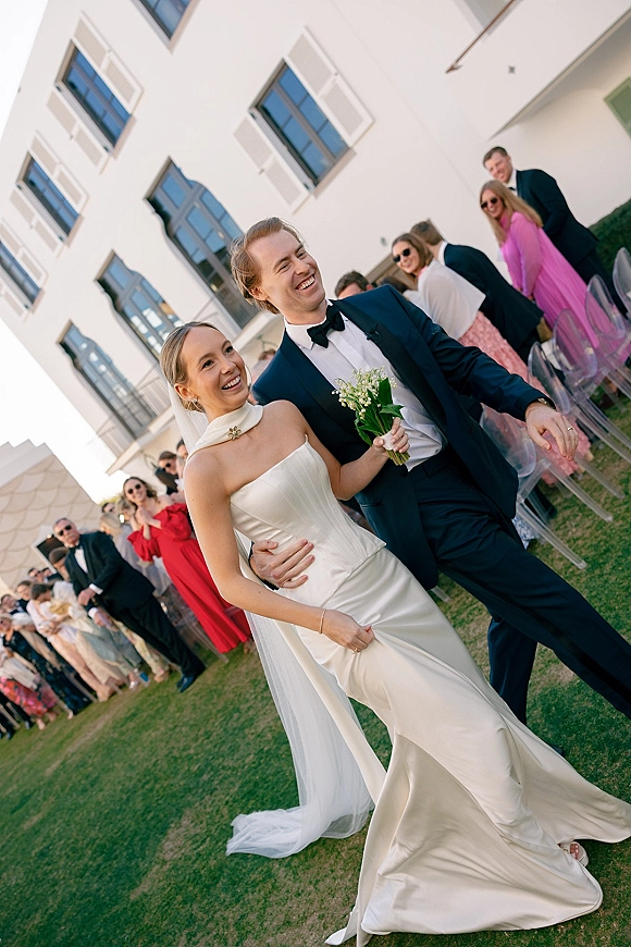 Recessional moment as bride and groom walk the aisle hand in hand, bride with bouquet and veil, guests cheering on outdoor lawn by white facade