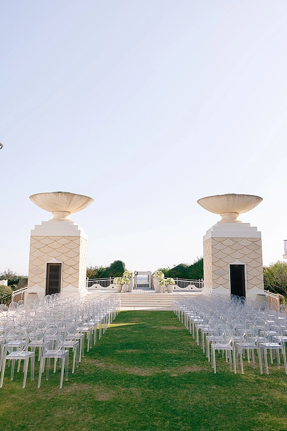 Ceremony setup for an outdoor wedding ceremony with clear acrylic chairs lining a white aisle runner, florals on stone steps before white towers