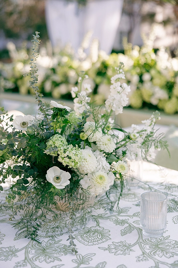 Wedding centerpiece with white flowers and greenery in a vase on a patterned tablecloth, with a ribbed glass votive in a garden setting