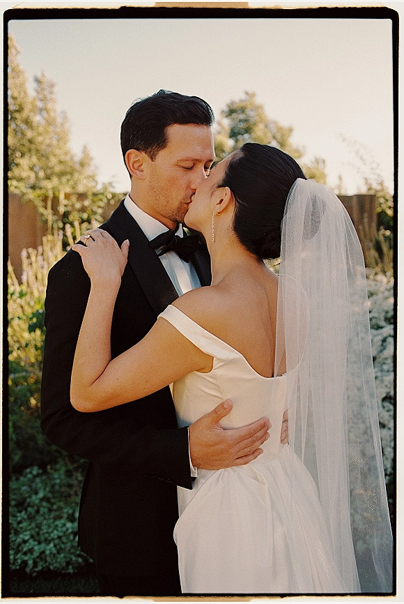Wedding kiss portrait of bride and groom kissing, her tulle veil flowing as he wears a black tuxedo against a garden fence backdrop