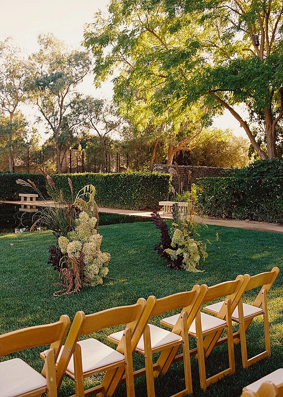 Ceremony setup with outdoor ceremony seating, wood folding chairs and hydrangea florals on a sunny lawn by trees and pond