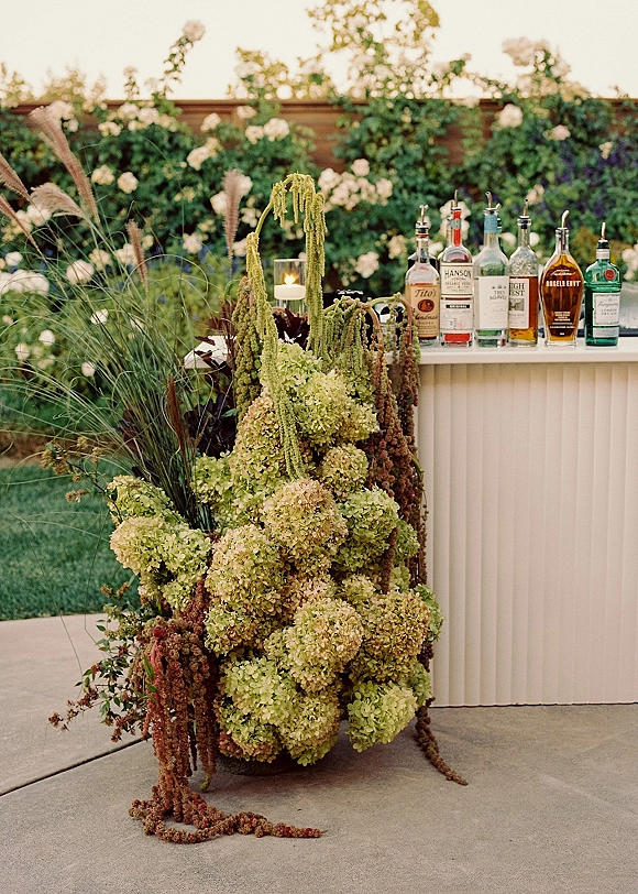 Wedding bar setup with outdoor wedding bar bottles on a white counter, framed by hydrangeas and hanging amaranthus in a garden patio setting