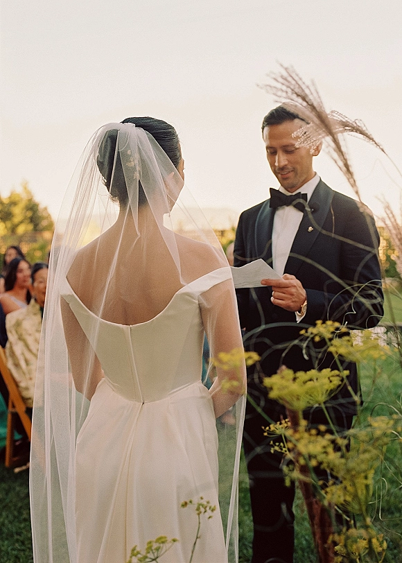 Wedding vows as groom reading vow paper beside bride in veil and off-the-shoulder dress at sunset outdoor garden ceremony with guests
