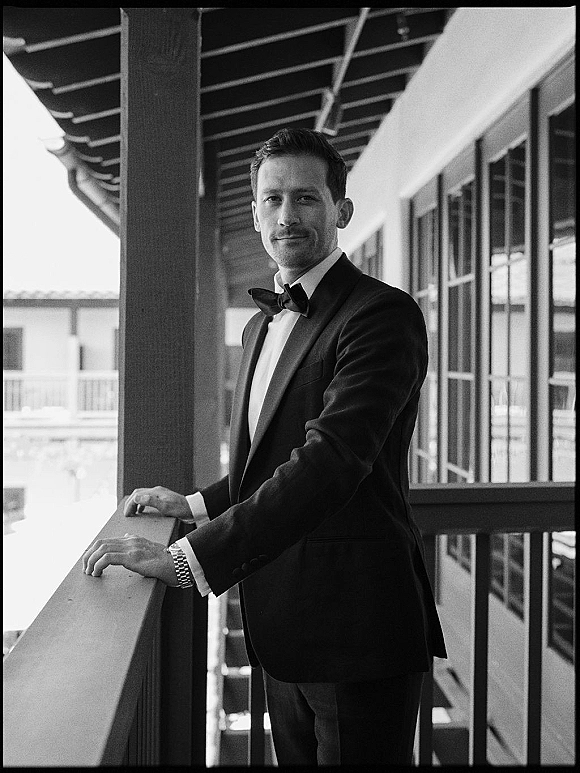 Groom portrait in a black tuxedo with bow tie and wristwatch, leaning on an outdoor balcony railing with columns and windows behind