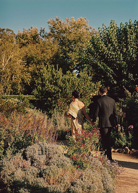 Couple walking away, bride and groom walking hand in hand with a gold sari and black suit along a garden path under blue sky