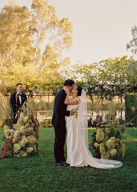 Wedding kiss as bride in veil and sleek gown kisses groom in black tux beneath hydrangea and pampas florals by a pond lawn