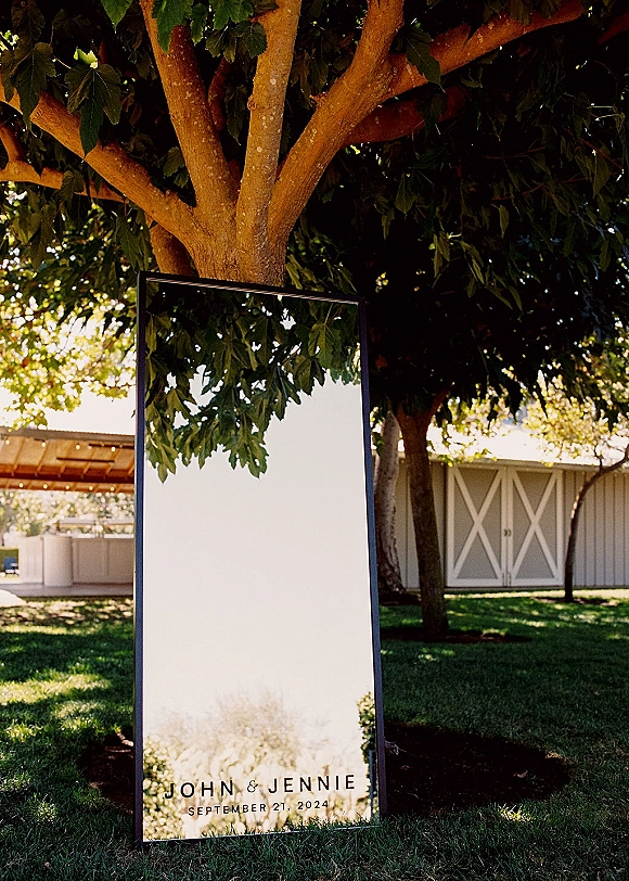 Wedding welcome sign, a framed mirror sign on an easel in an outdoor courtyard with barn doors, trees, and grass lawn behind