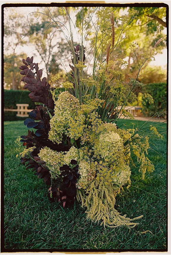 Wedding floral arrangement with hydrangea wedding centerpiece styling, greenery and tall grass stems on a garden lawn near trees and a bench